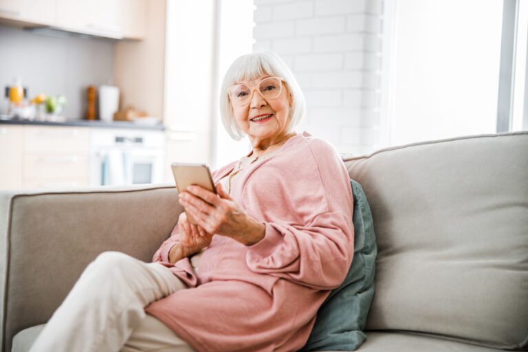 Uśmiechnięta starsza pani siedzi na kanapie i trzyma smartfona